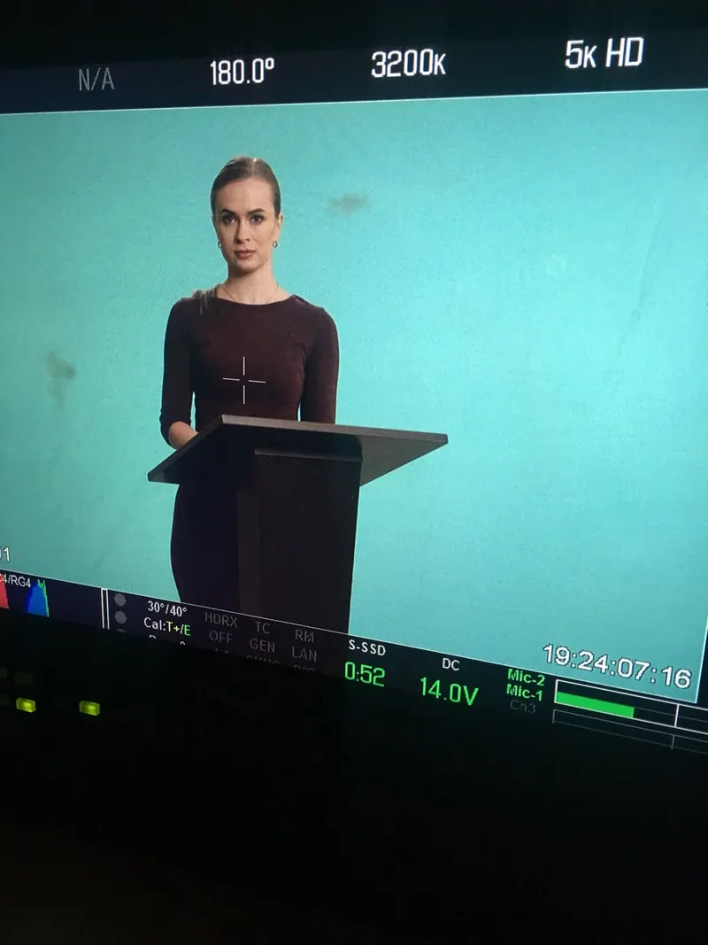 Krystyna at a podium in front of a green screen, seen on a camera monitor